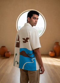 Man wearing a patterned shirt standing in a room with round window and vases.