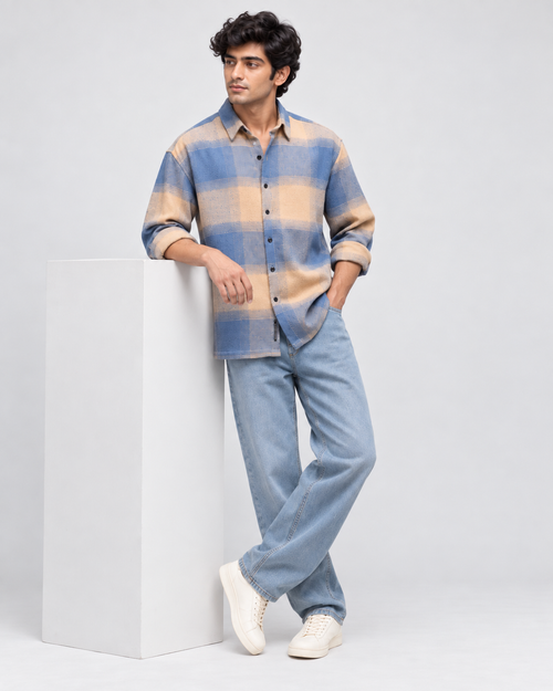 A man models a blue and beige plaid button-up shirt with light blue jeans and white sneakers, leaning against a white rectangular pedestal on a light gray studio background.