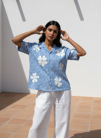 Woman wearing a blue checkered shirt with white floral patterns and white pants, standing against a light-colored wall.