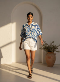 Woman in a blue and white patterned shirt and white shorts walking in a sunlit room with a vase of plants.