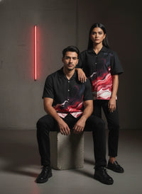 Two people wearing matching black and red patterned shirts sitting on a concrete block against a dark background.