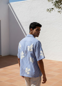 Man wearing a blue checkered shirt with floral patterns standing against a white wall.