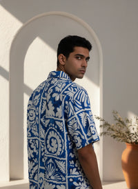 Man wearing a blue and white patterned shirt standing in a sunlit room with an archway.