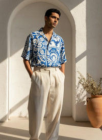 Man wearing a blue patterned shirt and white pants standing in a sunlit archway.