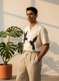 Man wearing a white shirt with black bird designs standing next to a potted plant against a light wall.