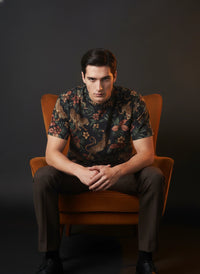 Man sitting in a brown chair wearing a patterned shirt against a dark background