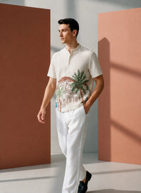 Man wearing a white shirt with a graphic design and white pants standing against a minimalistic background.