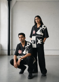 Two people wearing black outfits with white and pink patterns in a minimalistic setting.