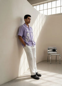 Man in a purple shirt leaning against a white wall with a chair in the background.