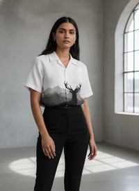 Woman wearing a white blouse with a deer graphic and black pants in a room with a large window.