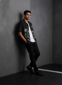Man leaning against a gray wall wearing a black shirt with white graphics and black pants.