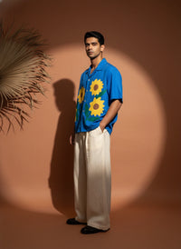 Man wearing a blue shirt with sunflower patterns and white pants standing against a brown background