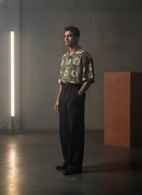 Man wearing a patterned shirt and black pants standing in a dimly lit room with concrete walls.