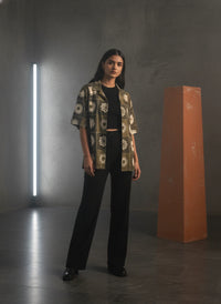 Woman in a patterned shirt and black pants standing in a dark room with a light vertical column and a red rectangular object.